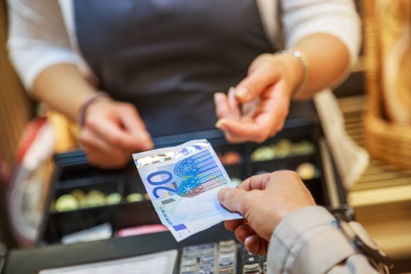 Close-up van een contante betaling bij een kassa. Een hand geeft een biljet van 20 euro aan de hand van een ondernemer, boven een open kassalade met munten en biljetten.