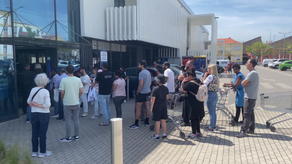 Een rij mensen met winkelwagens staat buiten te wachten voor de ingang van een supermarkt op een zonnige dag, met een drukke en onrustige sfeer.